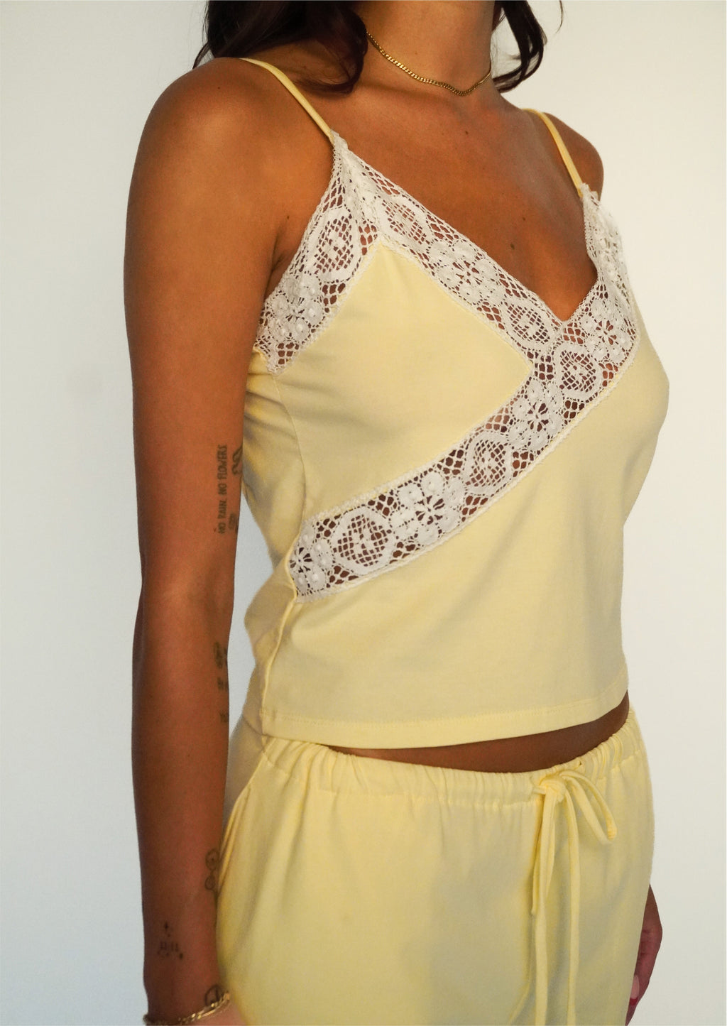 A woman wearing a pale yellow camisole top with white lace trim and matching pale yellow shorts against a plain light background.