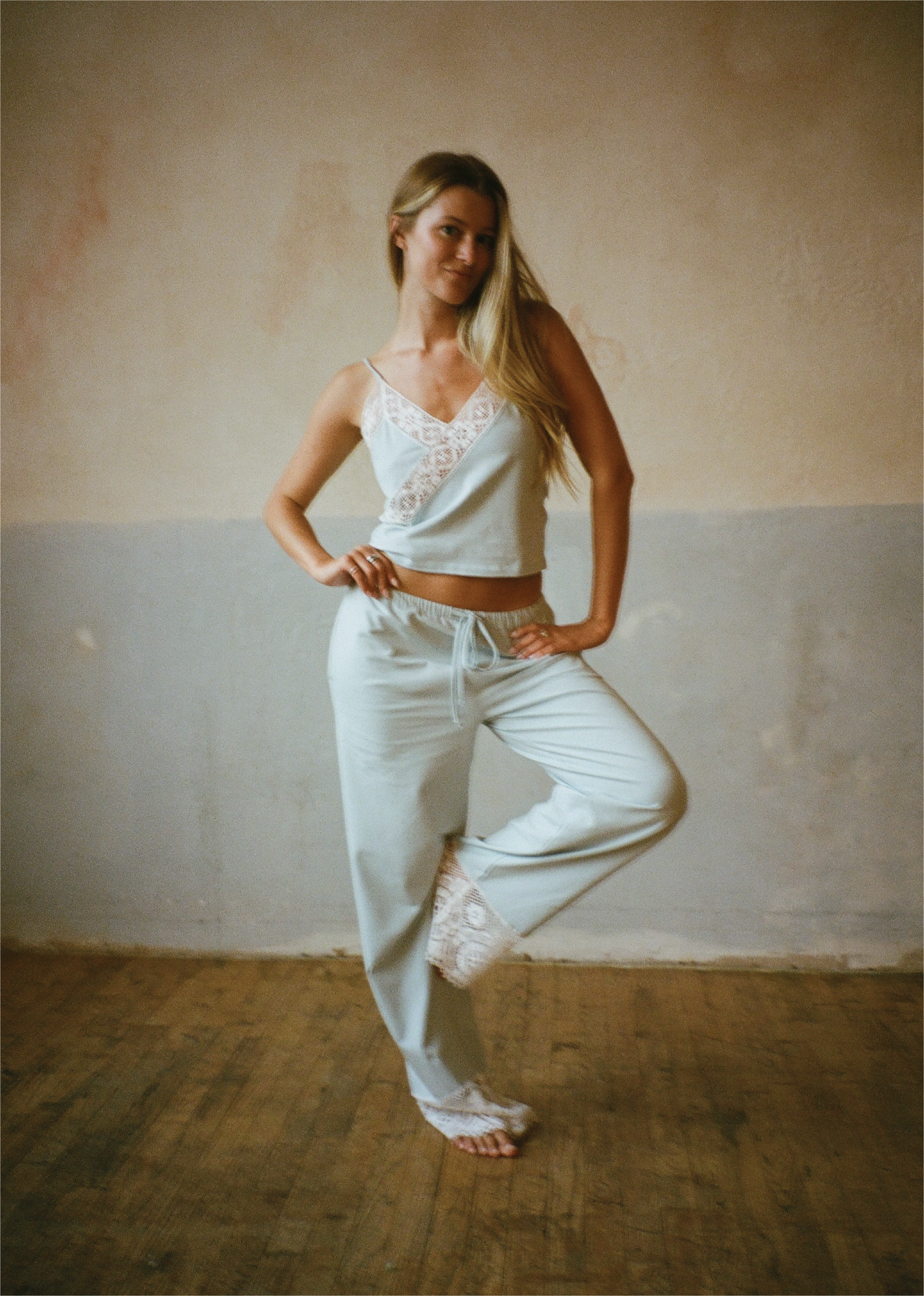 A woman modeling a light sage green lounge set consisting of a camisole top with white lace trim and matching sweatpants, standing in a rustic room with a textured plaster wall and wooden floor.