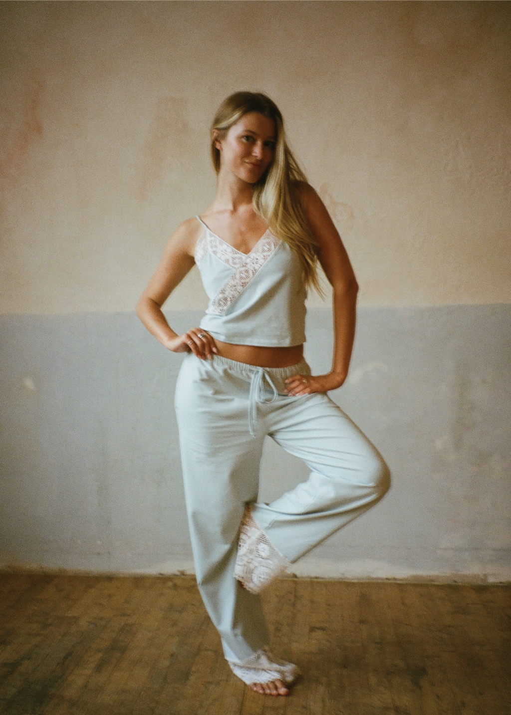A woman modeling a light blue lounge set consisting of a camisole top with white lace trim and matching sweatpants, standing in a rustic room with a textured plaster wall and wooden floor.