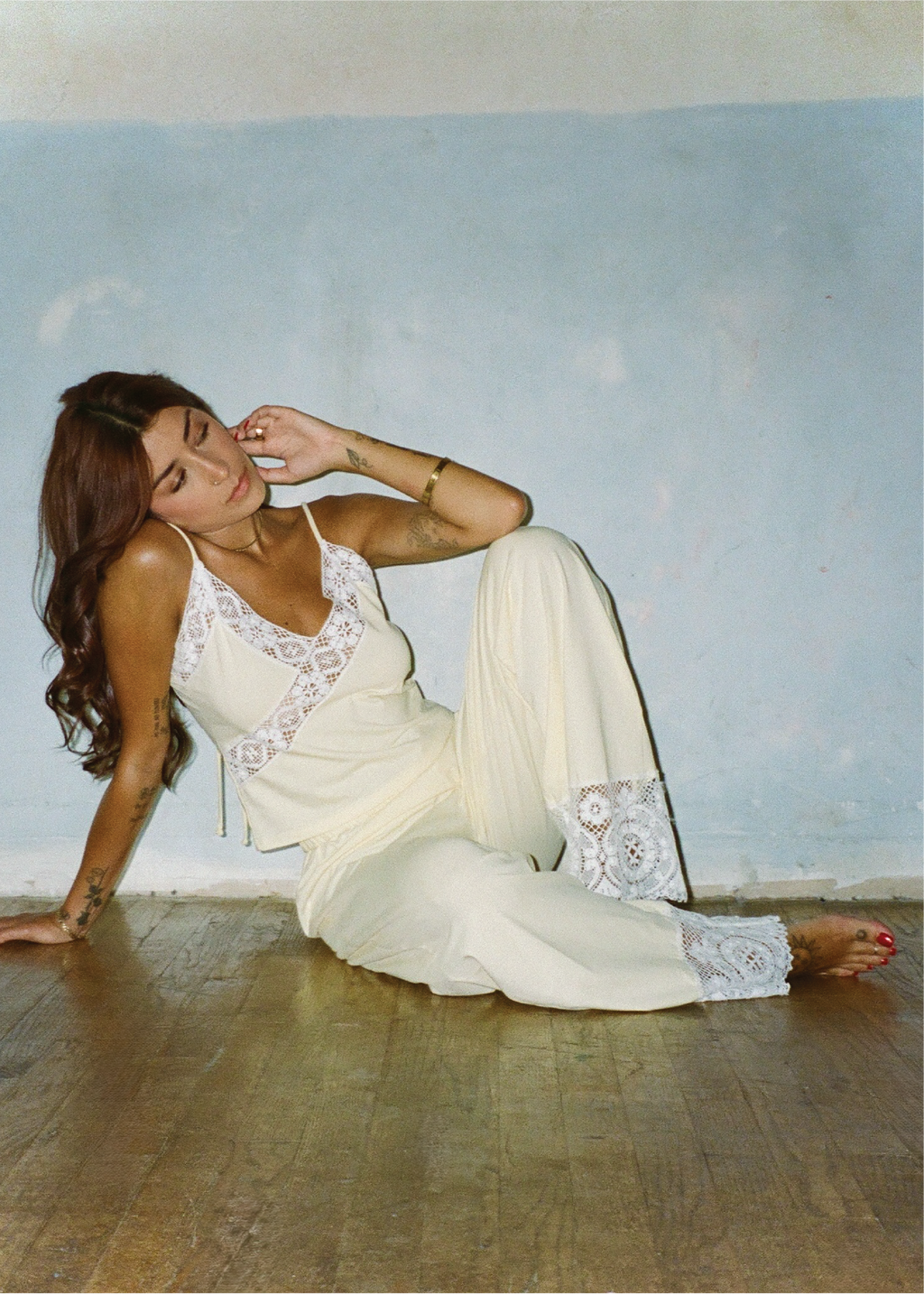 A woman reclining on a wooden floor against a light blue wall, wearing a cream-colored lace-trimmed camisole and matching wide-leg pants.