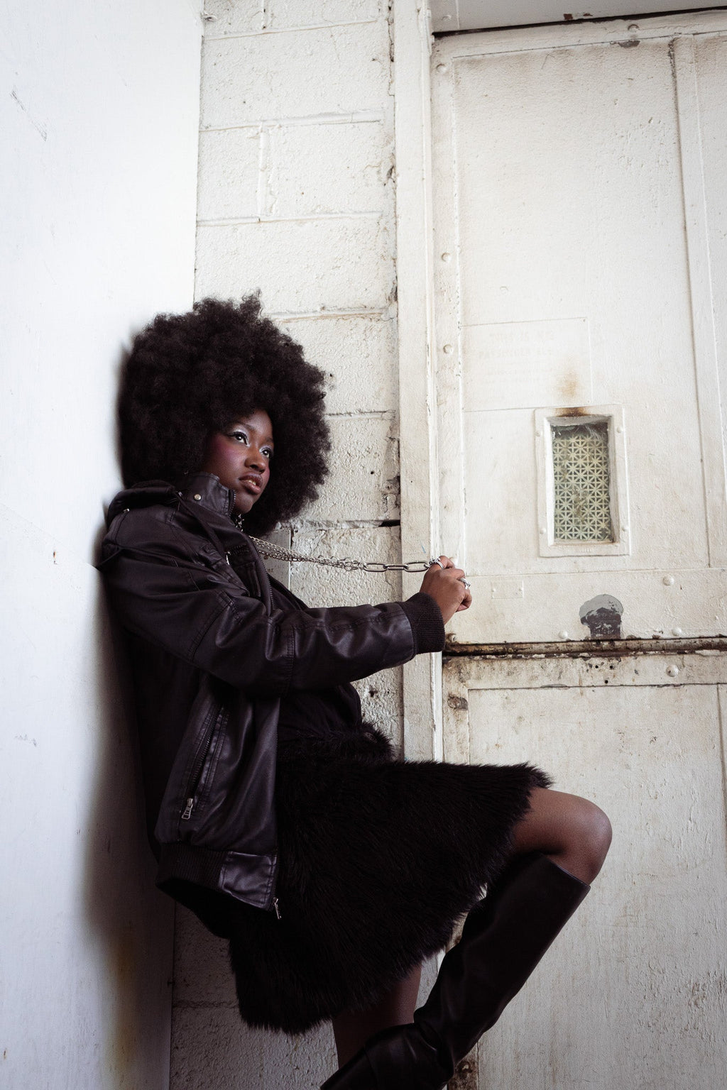 Woman with large afro wearing a black jacket, star earrings, and an aria chain with boots, chained to a door in a dimly lit room. 